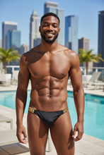 Load image into Gallery viewer, Man wearing black swim briefs with gold chains on the sides standing by a pool with city skyline in the background
